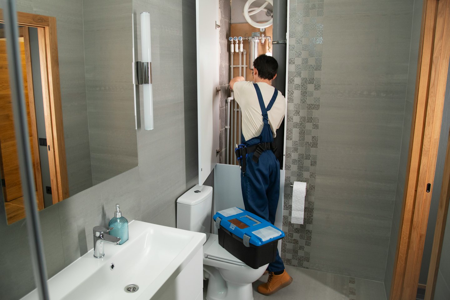Accueil Un plombier répare une canalisation derrière des toilettes dans une salle de bain carrelée de gris.